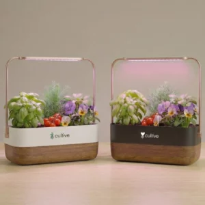 Deux potagers d'intérieur Cultive, modèles blanc et noir, posés côte à côte sur une table en bois avec leurs plantes éclairées